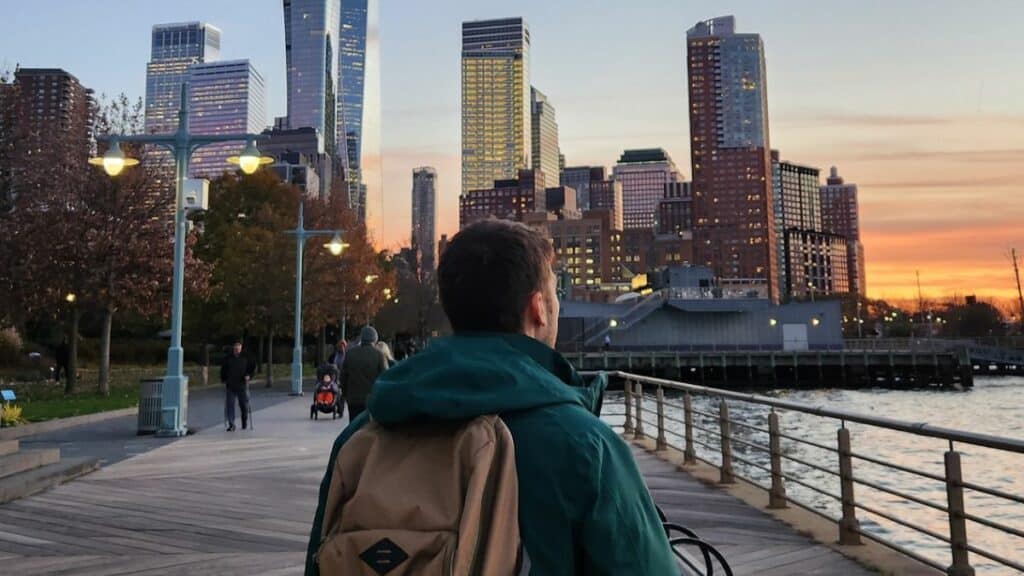 un homme de dos au coucher de soleil face aux immeubles de new york et en bord de mer