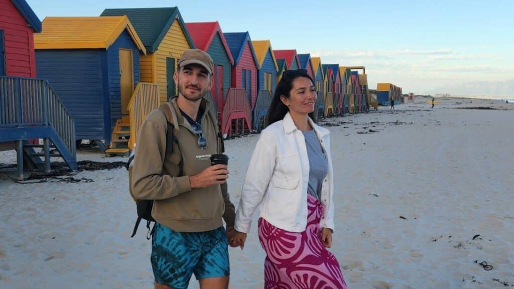un couple marchant sur une plage en afrique du sud, main dans la main devant des maisons colorées de Muizenberg